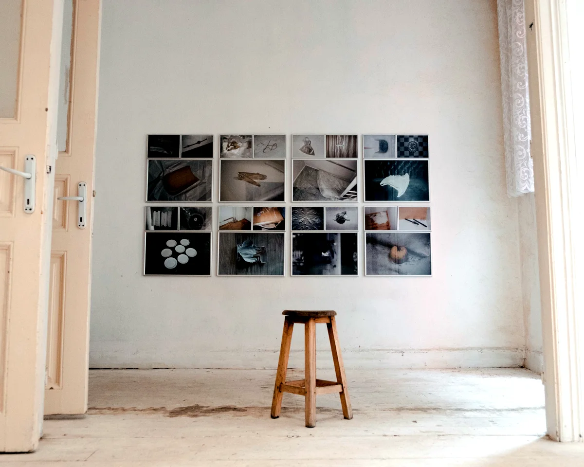 A grid of small works on a white wall with a wooden stool below it.