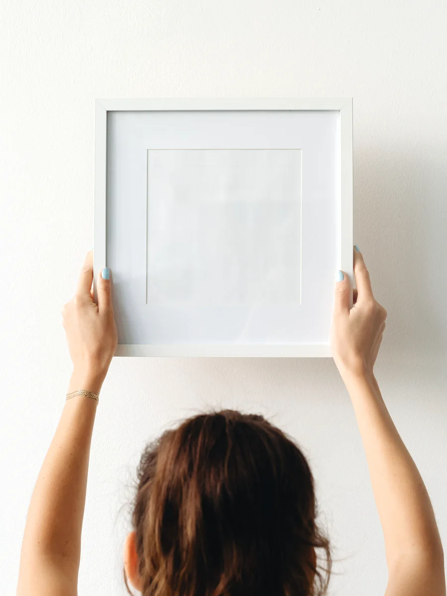 Two hands holding an empty frame flat against a white wall.